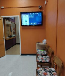 Hallway with chairs on the right and a tv screen on the wall.